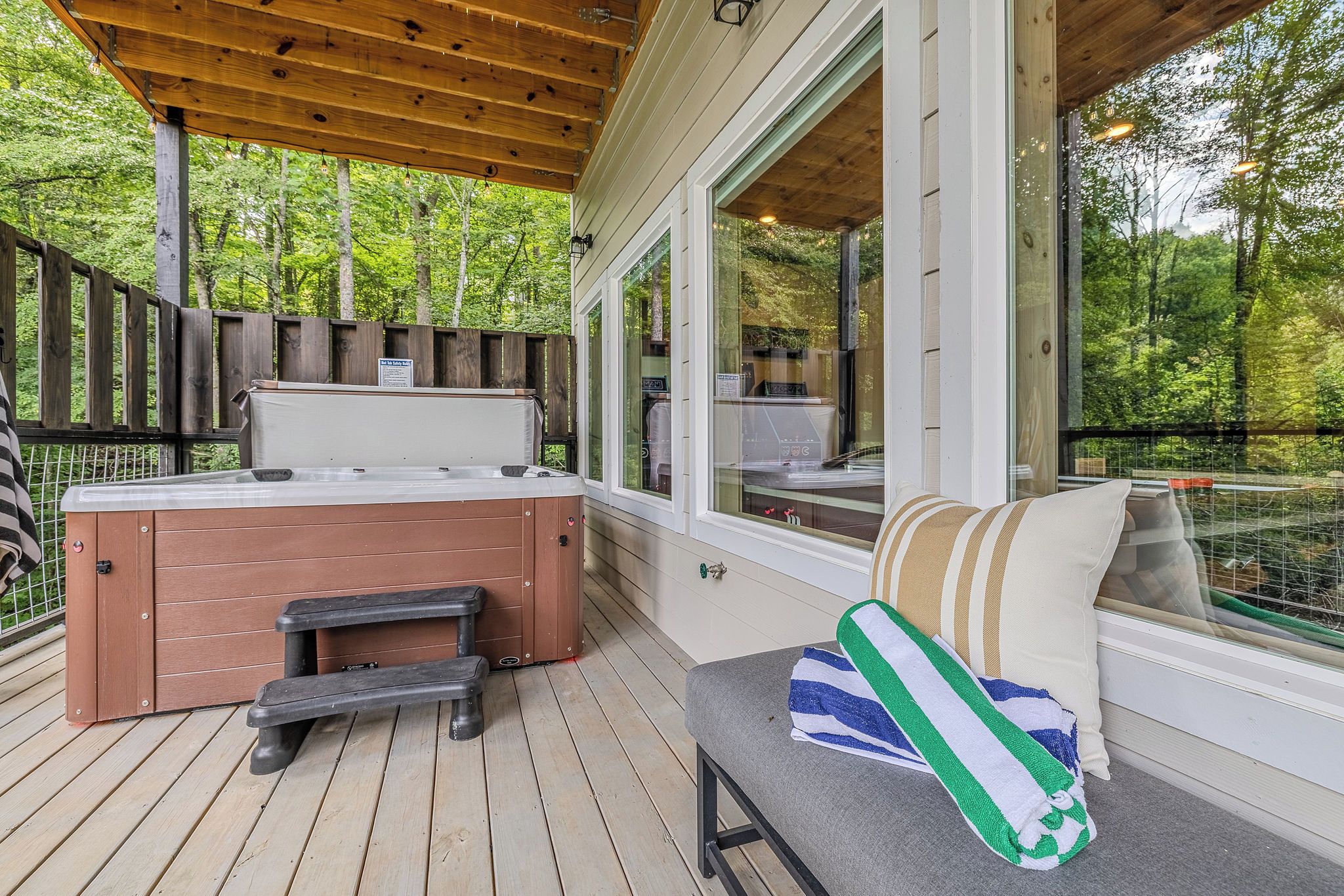 A wooden deck with a hot tub, a bench, and a cushioned seating area with a towel.