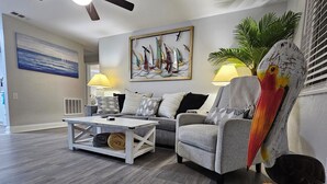 A living room with a grey sofa, a coffee table, and a painting of sailboats. There is a wooden bird sculpture and a lamp on a side table. - Seaside Serenity-Tranquil Beachside 2-bedroom home in beautiful Ormond-By-Sea (Ormond Beach)