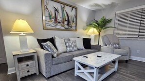 A living room with a grey sofa, two side tables with lamps, a coffee table, and a painting of sailboats. - Seaside Serenity-Tranquil Beachside 2-bedroom home in beautiful Ormond-By-Sea (Ormond Beach)