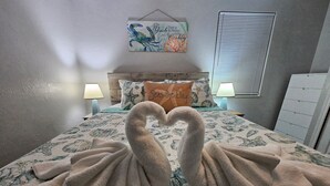 A bed with a heart-shaped towel arrangement, a wall with a seashore-themed painting, and a window with blinds. - Seaside Serenity-Tranquil Beachside 2-bedroom home in beautiful Ormond-By-Sea (Ormond Beach)