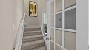 Una escalera con peldaños cubiertos de alfombra color beige, paredes blancas y una obra enmarcada en la parte superior.