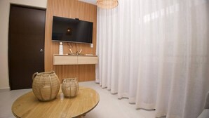 Un salon moderne avec un mur recouvert de panneaux en bois, un téléviseur à écran plat, une table ronde en verre agrémentée de deux vases en fibres naturelles et des rideaux blancs.