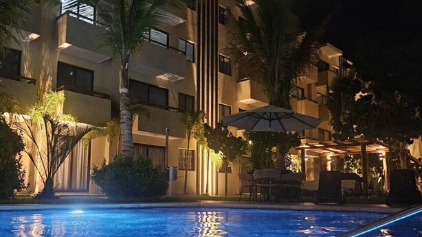 Une piscine éclairée pour la baignade nocturne, avec un bâtiment en arrière-plan, des palmiers et des espaces de détente en plein air.