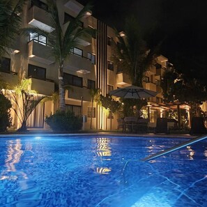 Ein Schwimmbeckenbereich bei Nacht mit einem Gebäude im Hintergrund, Palmen und Außenmöbeln.