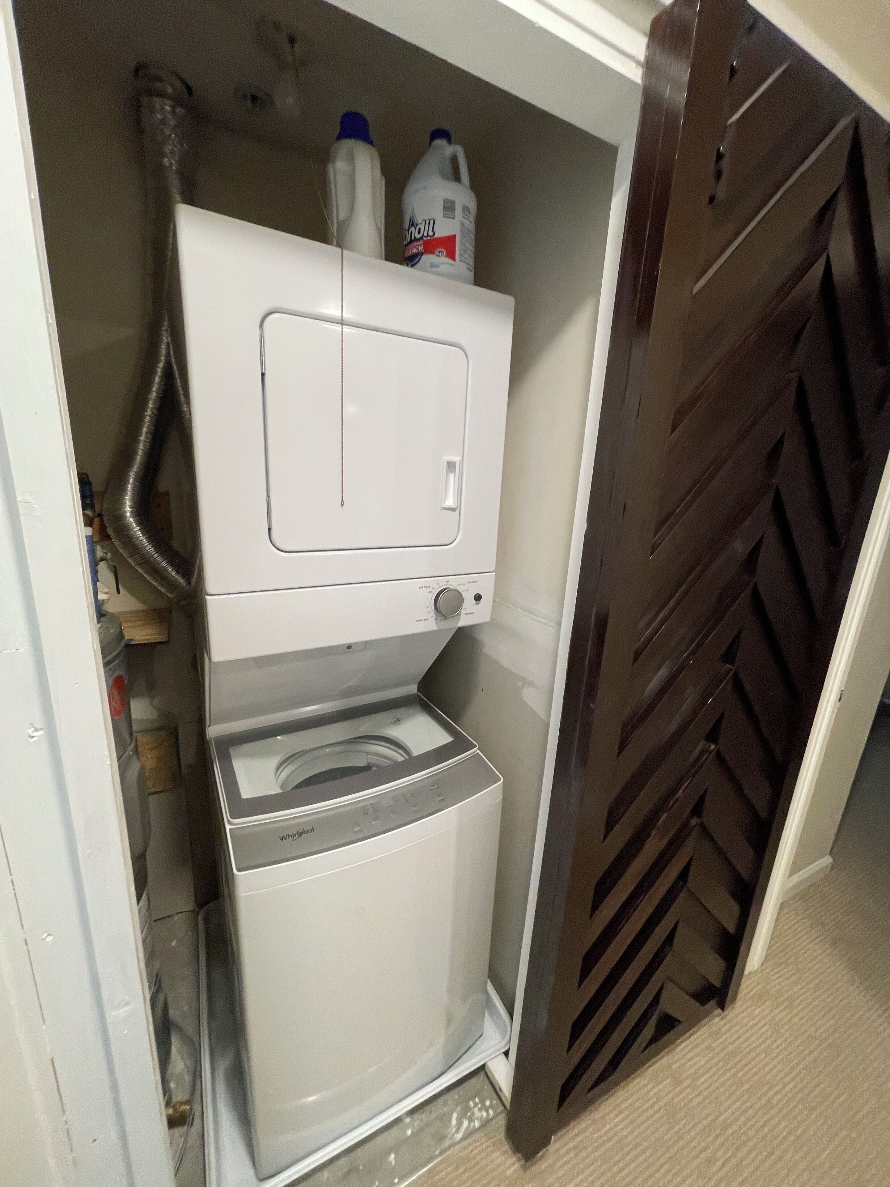 A compact, white washing machine with a front-loading door, located in a small utility space with a wooden door and a metal vent.
