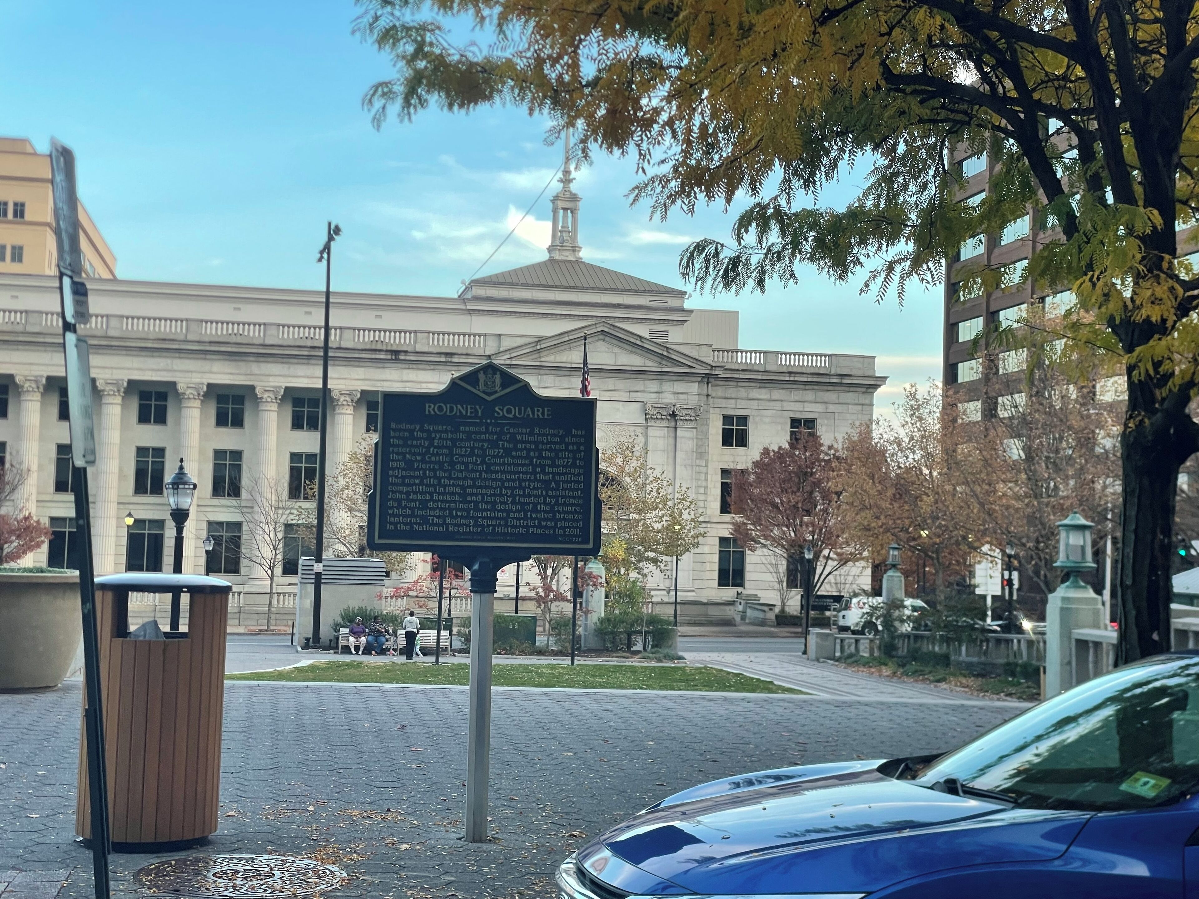 A historical marker sign in front of a classical building with columns, a dome, and a statue on top.