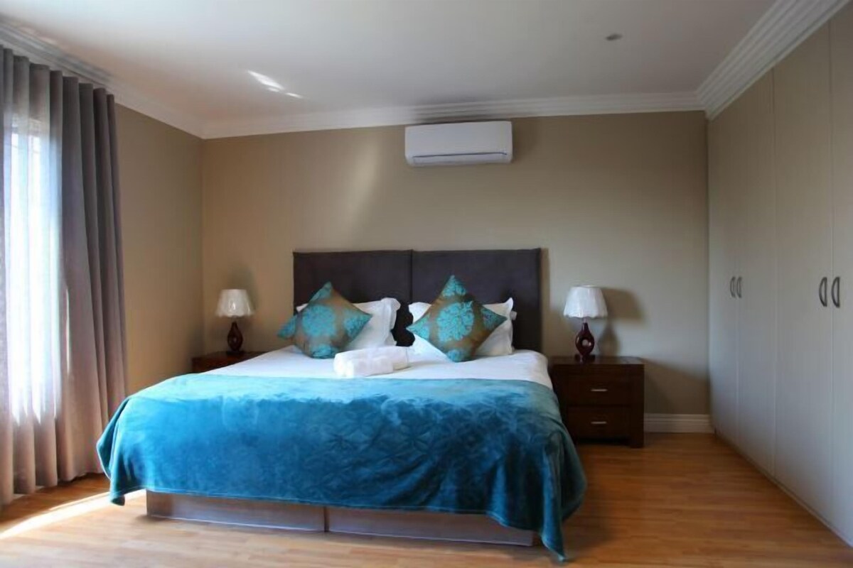 A bedroom with a bed, two bedside tables, a nightstand, a window with curtains, and an air conditioning unit on the wall.