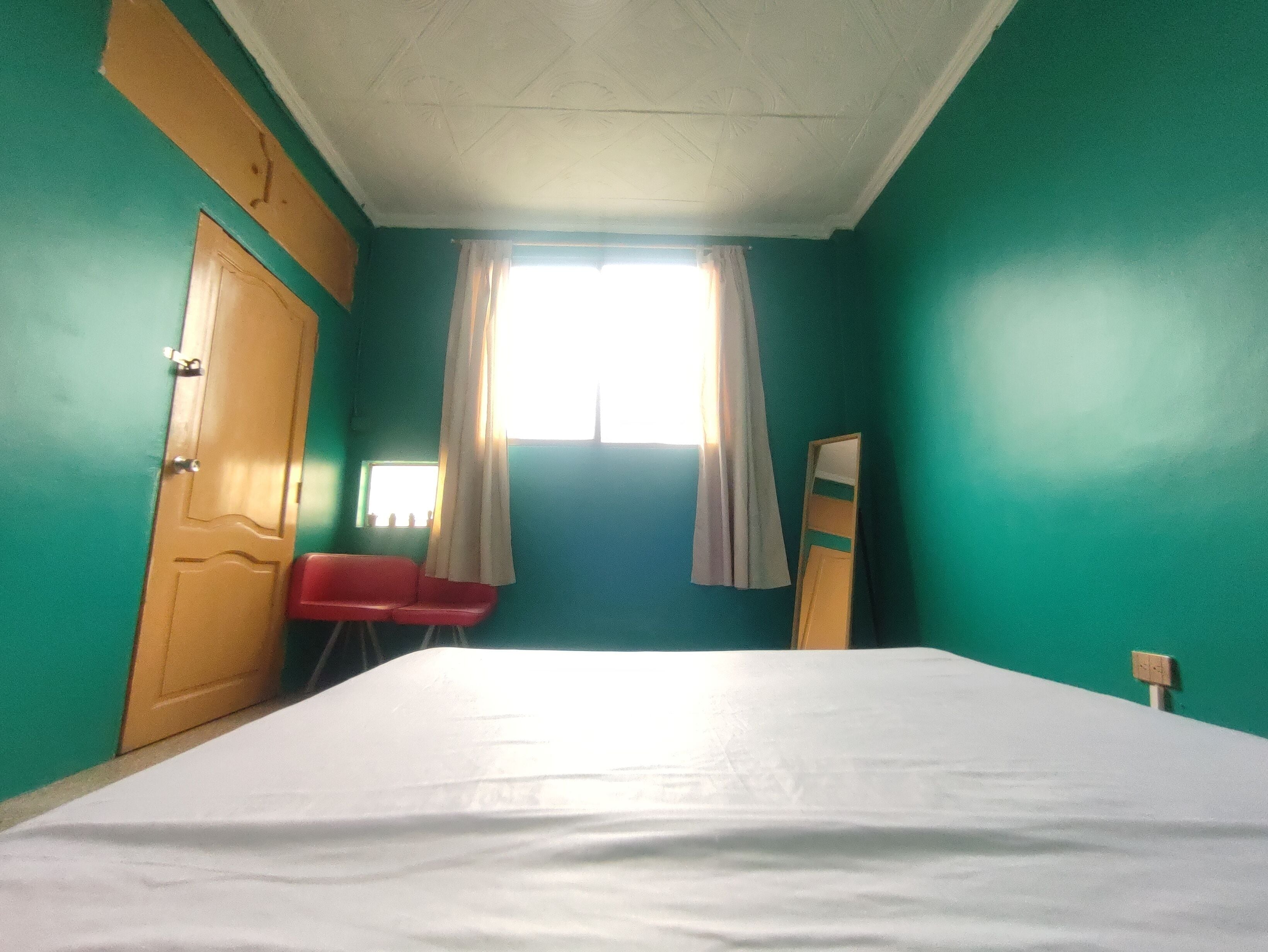 A room with teal walls, a wooden door, a bed with a white sheet, a red chair, and a window with curtains.