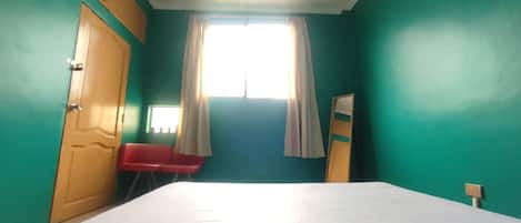 A room with teal walls, a wooden door, a bed with a white sheet, a red chair, and a window with curtains.