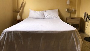 A neatly made bed with white linens in a room with yellow walls, a small shelf above the bed, a bedside table with a lamp, and a standing fan. - Pachamac House (Guayaquil)