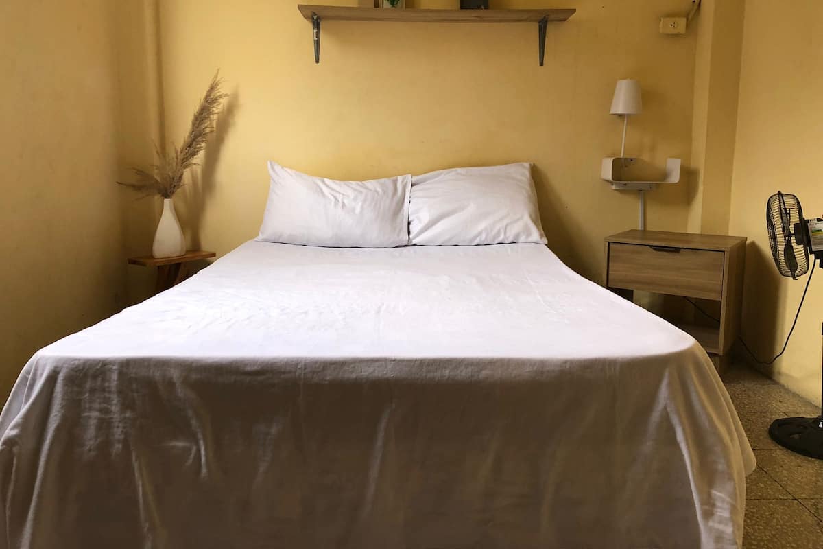 Egy rendezett ágy fehér ágyneművel, sárga falú szobában, az ágy felett egy kis polc, az ágy mellett egy éjjeliszemly és egy állóventilátor található.
