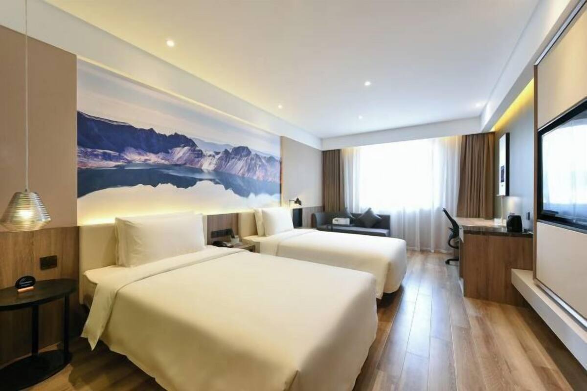 A hotel room with two beds, a desk, a large window, and a mountain landscape mural.