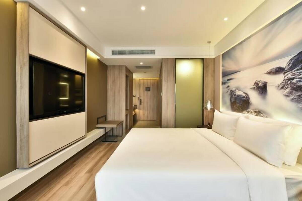 A modern hotel room with a large bed, a flat-screen TV, and a painting of a mountain landscape.