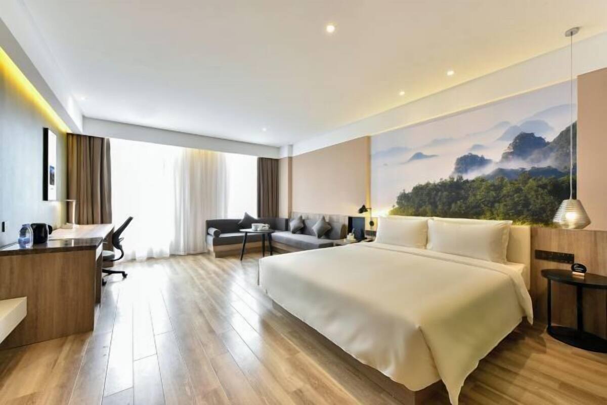 A modern hotel room with a large bed, a desk, a sofa, and a view of a mountainous landscape.