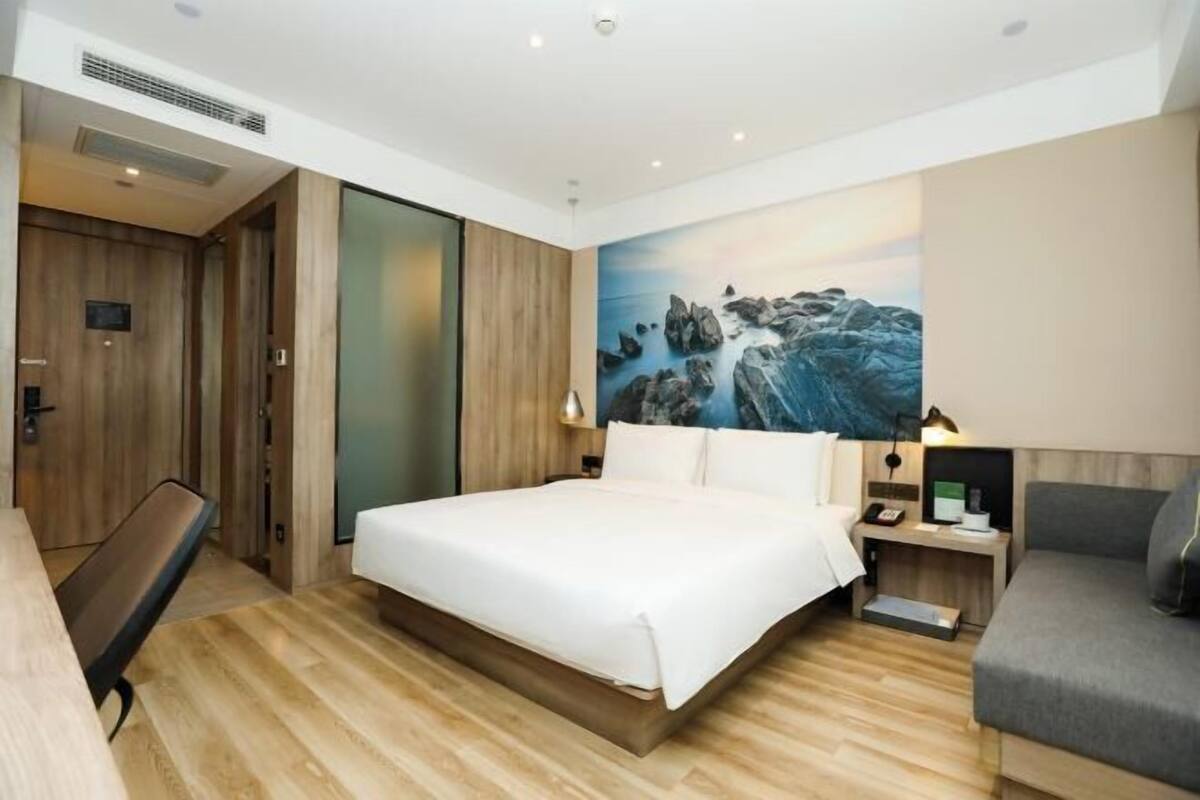 A modern hotel room with a wooden floor, a large bed, a desk with a chair, and a painting of rocks on the wall.