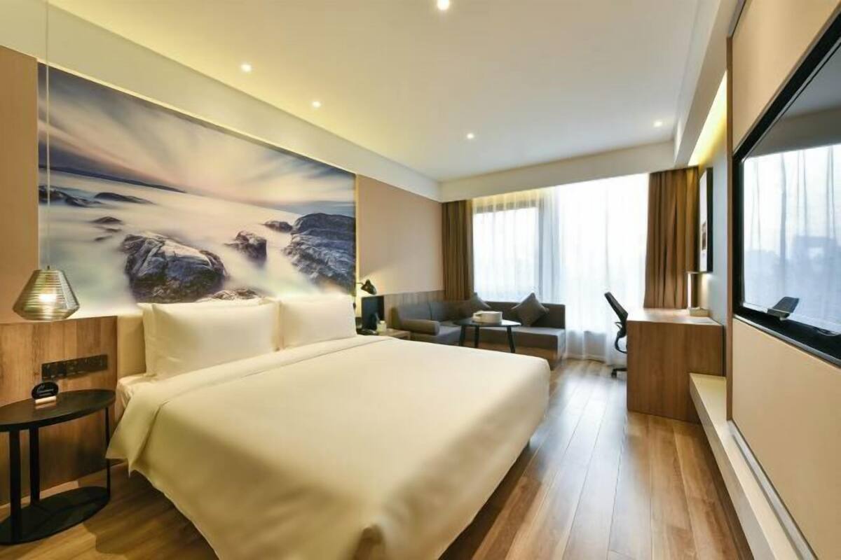 A modern hotel room with a large bed, a desk, a sofa, and a large window.