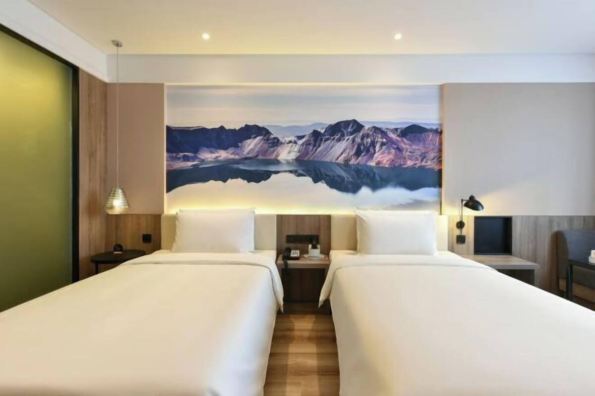 A hotel room with two beds, a large mountain painting on the wall, a wooden floor, and a nightstand with a lamp.