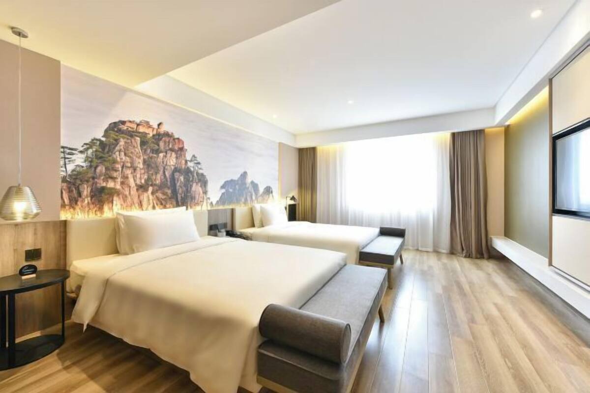 A hotel room with two beds, a large window with curtains, a wall-mounted TV, and a wooden floor.