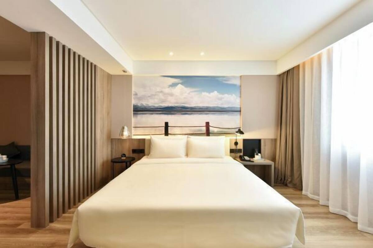 A hotel room with a large bed, a painting of a landscape, a desk with a lamp, and curtains on the windows.