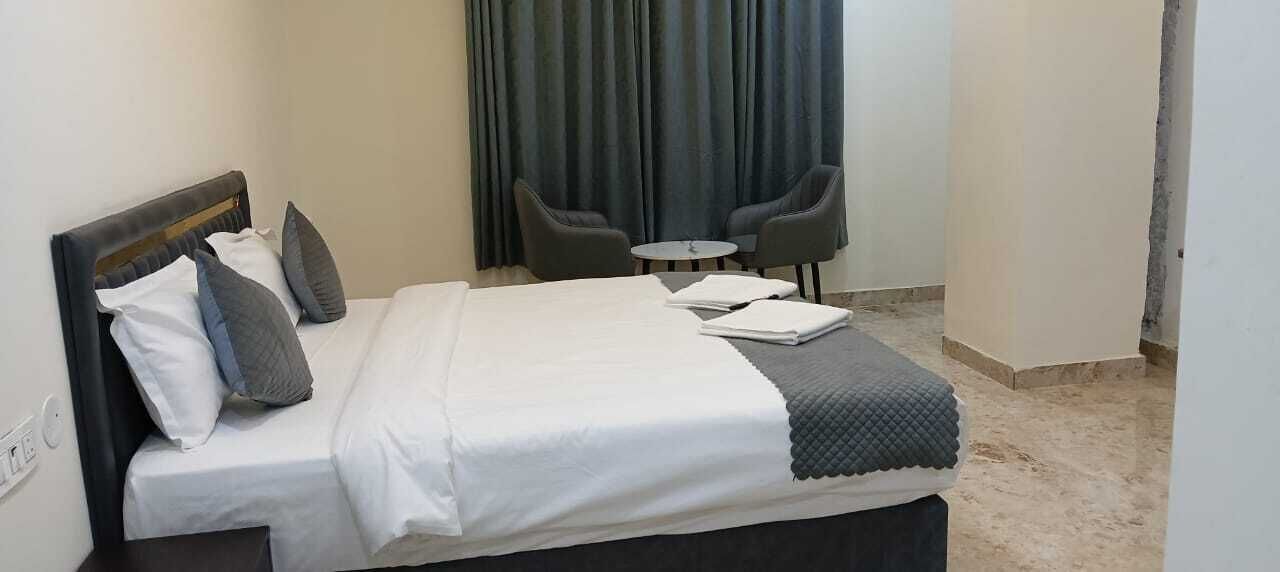 A neatly made bed with white and gray bedding, a small round table with two chairs, and a dark curtain in the background.