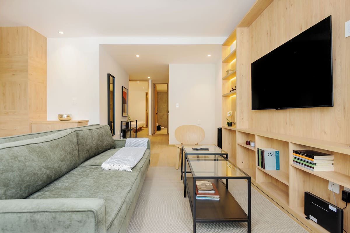 A modern living room with a grey sofa, a glass coffee table, a flat-screen TV mounted on a wooden wall, and a bookshelf with books.