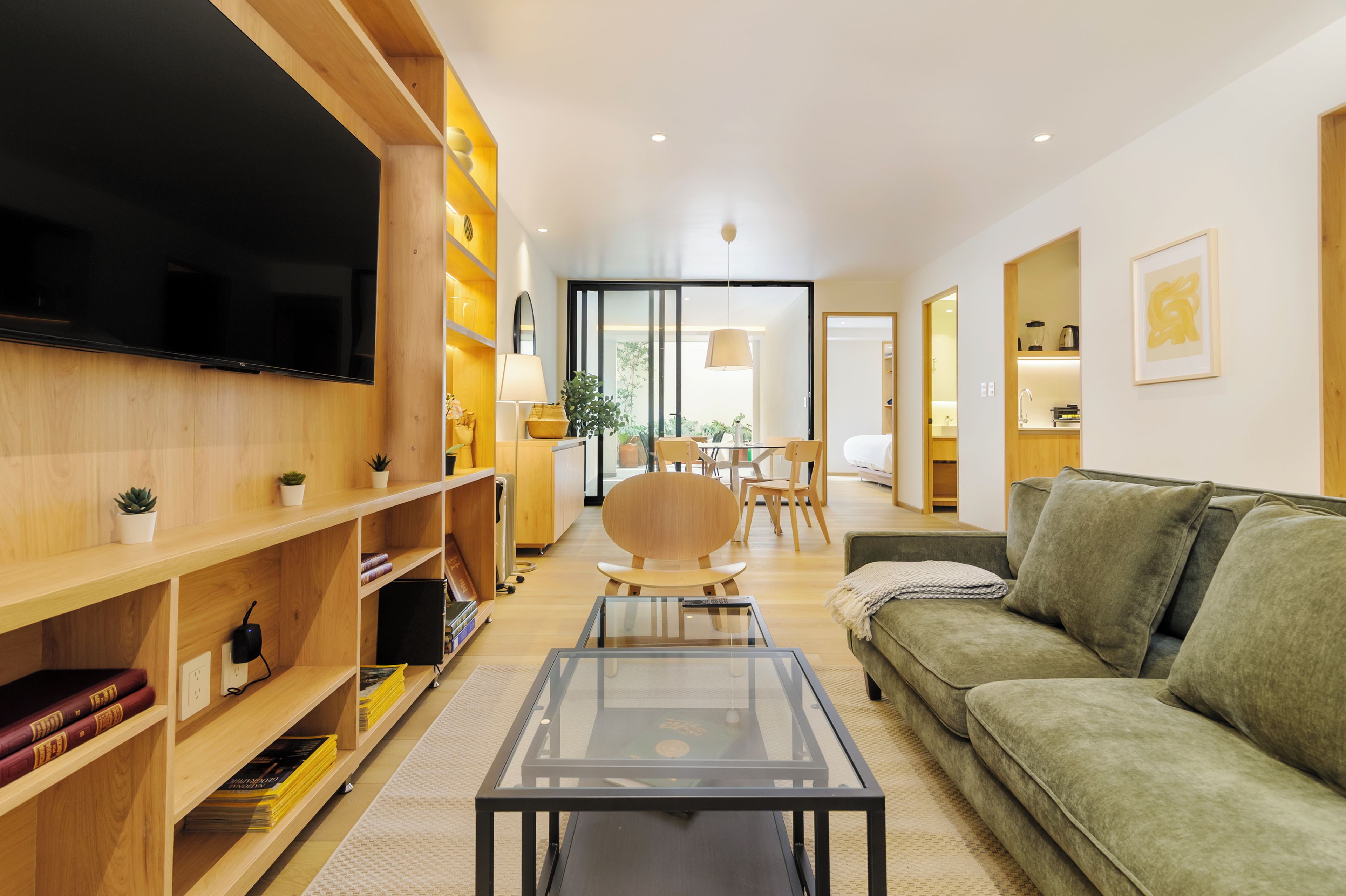 A modern living room with a flat-screen TV, wooden shelving, a green sofa, and a dining area with a table and chairs.
