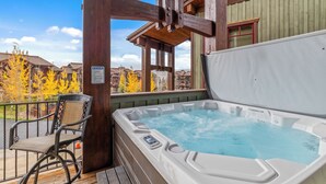 Un jacuzzi en una terraza de madera con una silla, una pérgola de madera y vistas a una zona residencial con árboles otoñales.