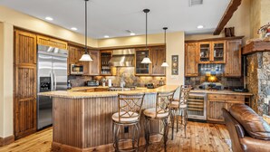 Cocina con armarios de madera, un refrigerador de acero inoxidable, una isla central con taburetes de bar e iluminación colgante.