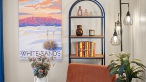 Ein gemütliches Zimmer mit einem weichen orangefarbenen Sessel, einem Bücherregal mit Büchern und Dekoration, einem gerahmten Kunstwerk mit Wüstenlandschaft und einer hohen Stehlampe.