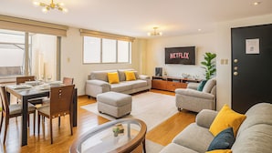 A modern living room with a dining area, a grey sofa with yellow cushions, a flat-screen TV on the wall, and a black door with a QR code. - Esmeralda Shelter - Jacuzzi (Cuenca)
