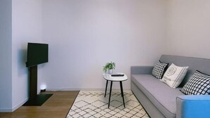 A living room with a grey sofa, a small round table with a plant, a flat-screen TV on a stand, and a patterned rug. - Green&Tree (Osaka)