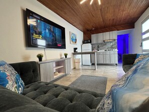 Un soggiorno moderno con una TV a schermo piatto, divani grigi, un soffitto in legno e un angolo cottura con pensili grigi e uno schienale in marmo.