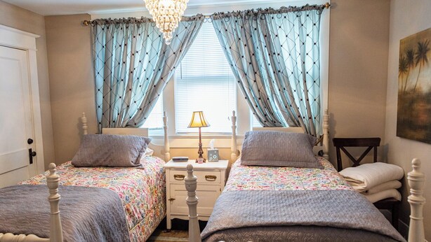 A bedroom with two beds, a chandelier, a bedside table with a lamp, and a painting on the wall. - Guest House of Kentucky (2 Singles - 2nd Floor) (Lancaster)
