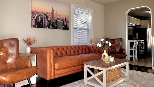 A modern living room with a brown leather sofa, a wooden coffee table, and a cityscape painting on the wall. - Cozy Home in Madison Heights (Madison Heights)