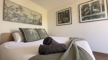 Ein ordentlich bezogenes Bett mit einer weißen Tagesdecke, zwei Kissen mit Palmenmotiven, einem gefalteten dunklen Handtuch und einer grünen Strickdecke.