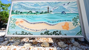 A colorful mural with text and illustrations of a beach scene, including a lighthouse, palm trees, and a turtle. - Lovely king room in 8 unit Inn in tranquil Sanibel with bikes, beach gear... (Sanibel)