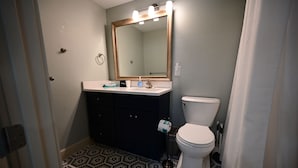 A bathroom with a white toilet, a dark-colored vanity, a large mirror with lights, and a patterned floor. - Lovely king room in 8 unit Inn in tranquil Sanibel with bikes, beach gear... (Sanibel)