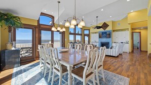 A spacious dining area with a large wooden table and white chairs, a kitchen in the background, and a view of the ocean through large windows. - Bella Vista II | 3,144 ft to the beach | Private Pool and Hot Tub (Corolla)