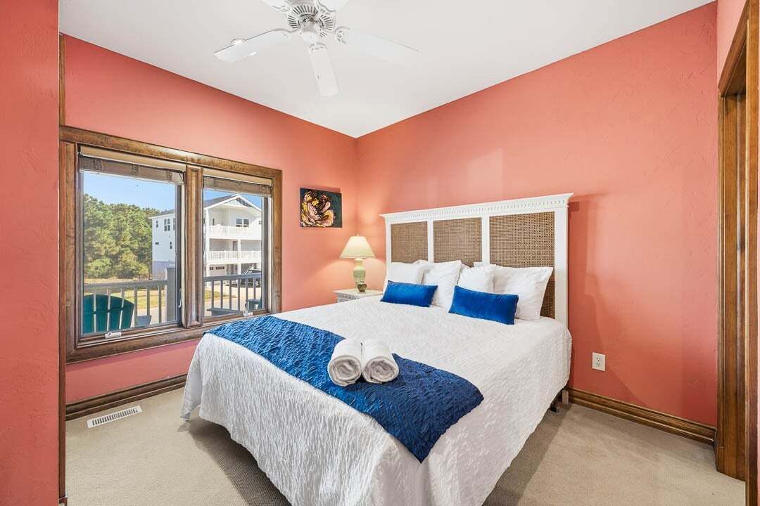Een slaapkamer met een groot bed, een raam met uitzicht op een huis, een plafondventilator en een nachtkastje met een lamp.