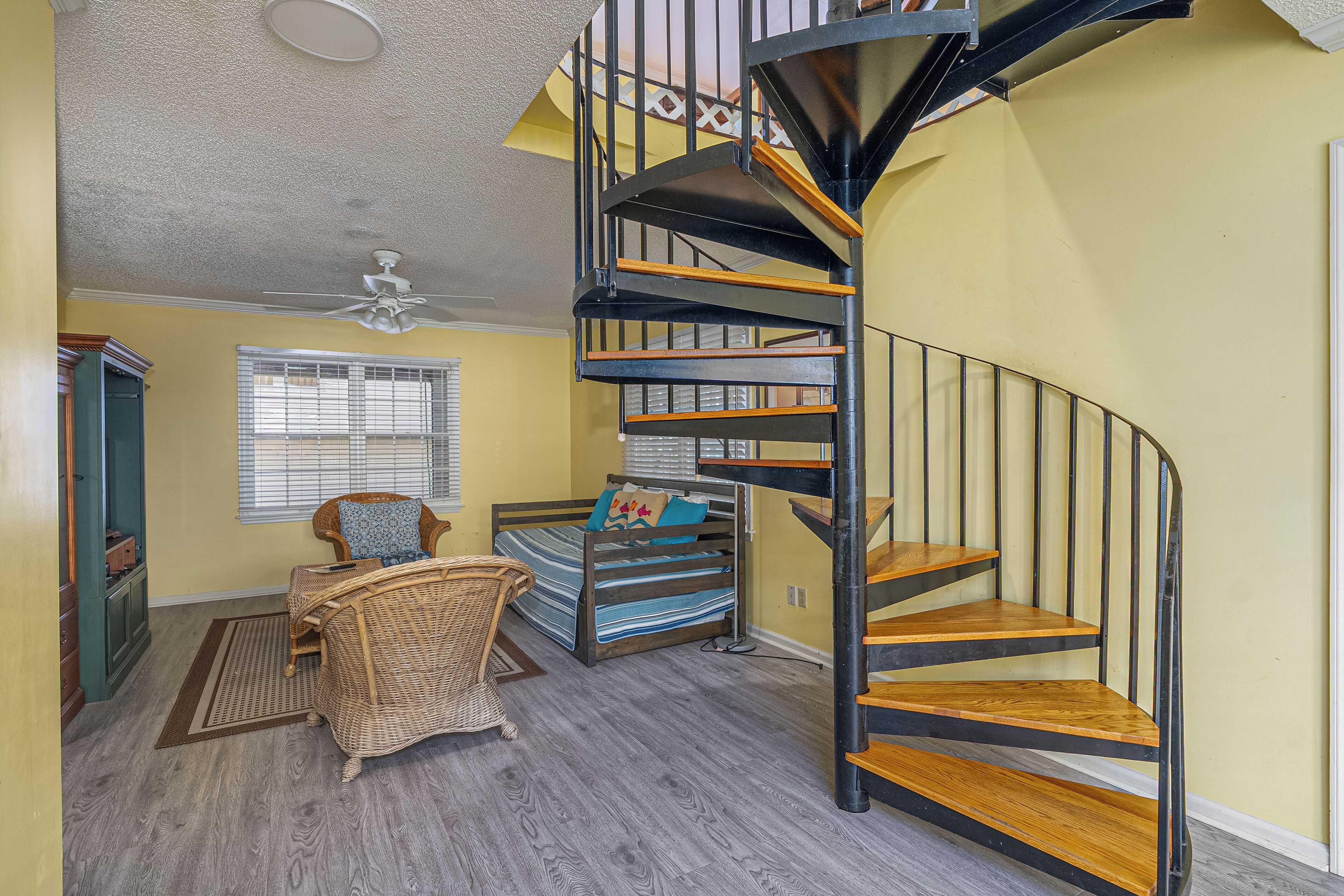 A room with a spiral staircase, a bed with a striped bedspread, a wicker chair, and a window with blinds.