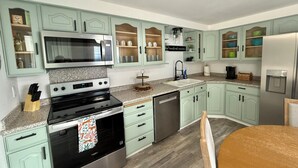 Une cuisine avec des armoires de couleur turquoise, un four à micro-ondes, un four et un lave-vaisselle en acier inoxydable. On y trouve un comptoir en granit, une table à manger en bois et un évier avec un robinet.