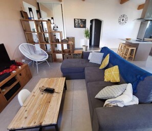 Ein modernes Wohnzimmer mit grauem Sofa, Holztisch und weißem Sessel. Ein Küchenbereich mit Herd und Spüle ist ebenfalls zu sehen.