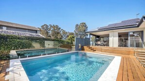 A swimming pool with a wooden deck, a covered patio, and a house with a solar panel roof.