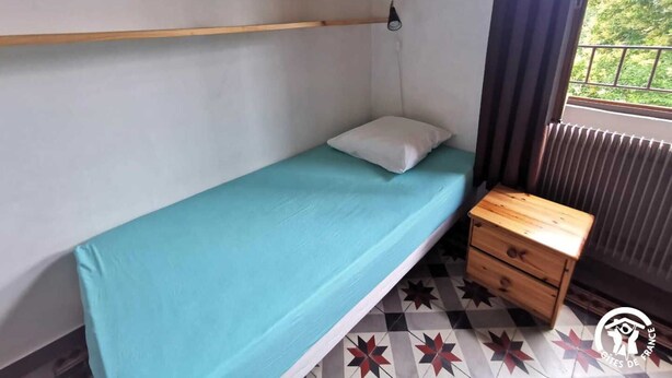 Ein Zimmer mit einem Einzelbett, einem dunkelblauen Bett, einem Nachttisch aus Holz und einem Fenster mit Blick auf Grün.
