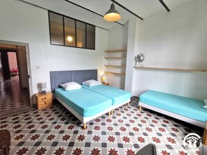 Une chambre avec un lit, une table de chevet, une lampe, un ventilateur et une fenêtre avec des stores.