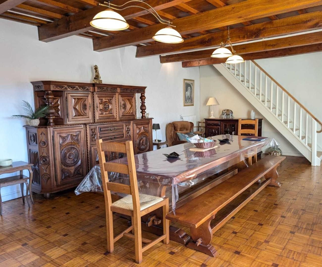 Un vaste espace dédié à la salle à manger, avec une table en bois, des chaises et un grand meuble. On aperçoit un escalier au fond.