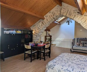 Un dormitorio ático acogedor con un arco de piedra, techo de madera, cama con colcha floral, una mesa de comedor con sillas y un armario negro con varios objetos.