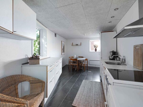 A compact kitchen with a dining area, white cabinetry, a wicker chair, and a small dining table. - 5 Star Holiday Home in Svaneke (Svaneke)