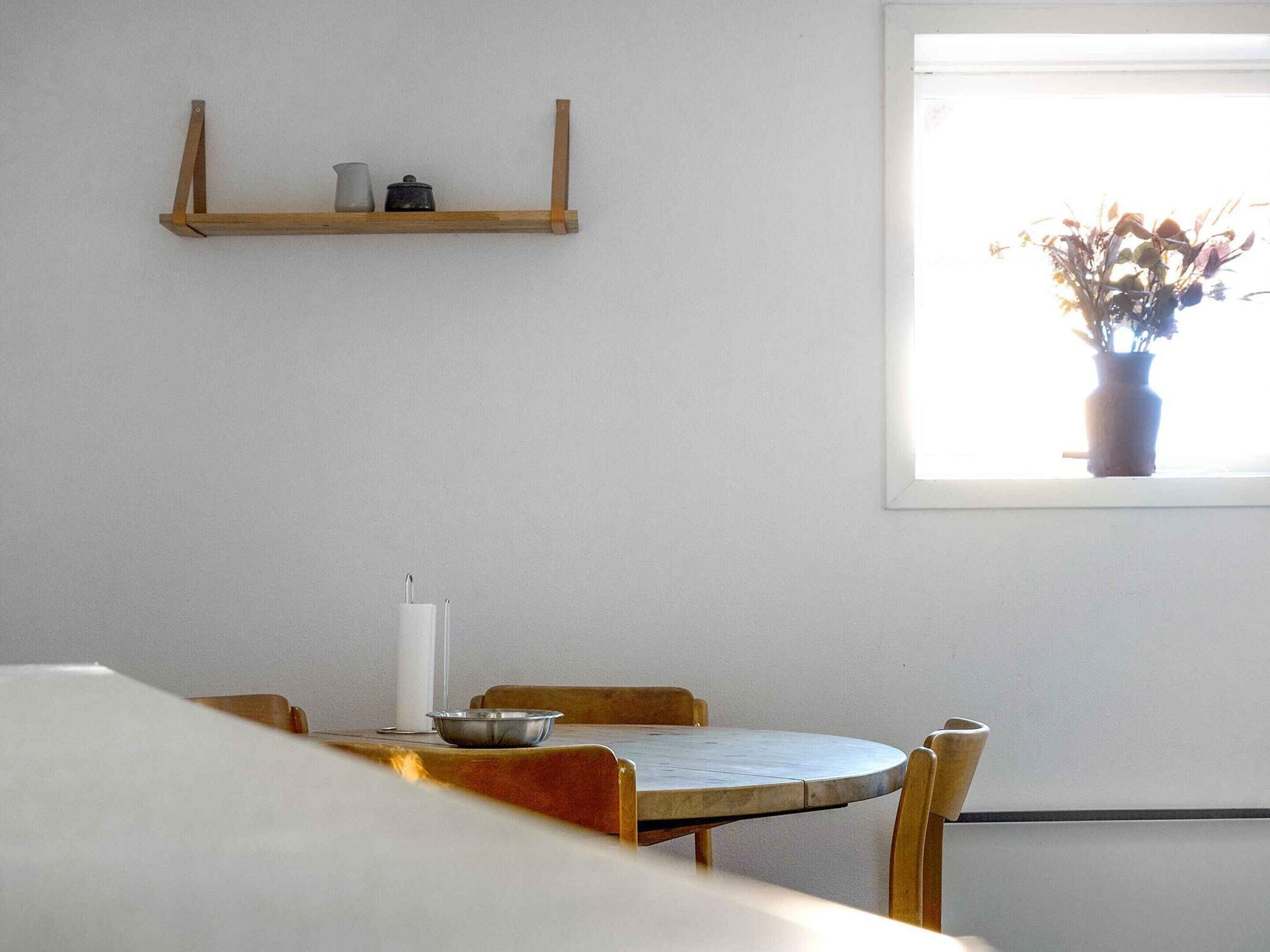 Un coin repas comprenant une table ronde, des chaises en bois, une petite étagère avec des ustensiles de cuisine et une fenêtre agrémentée d’une plante.