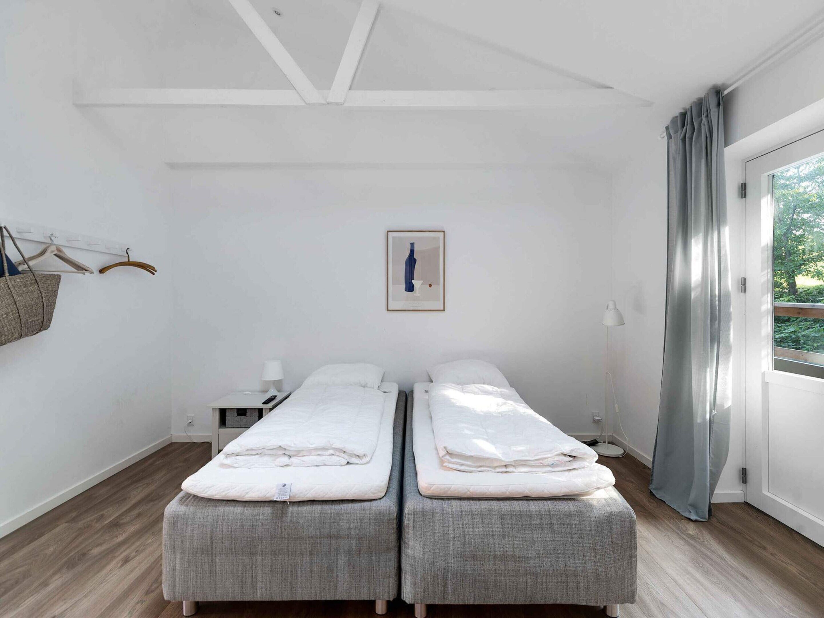 A twin room with two beds, a bedside table with a lamp, a hanging bag, and a window with curtains.